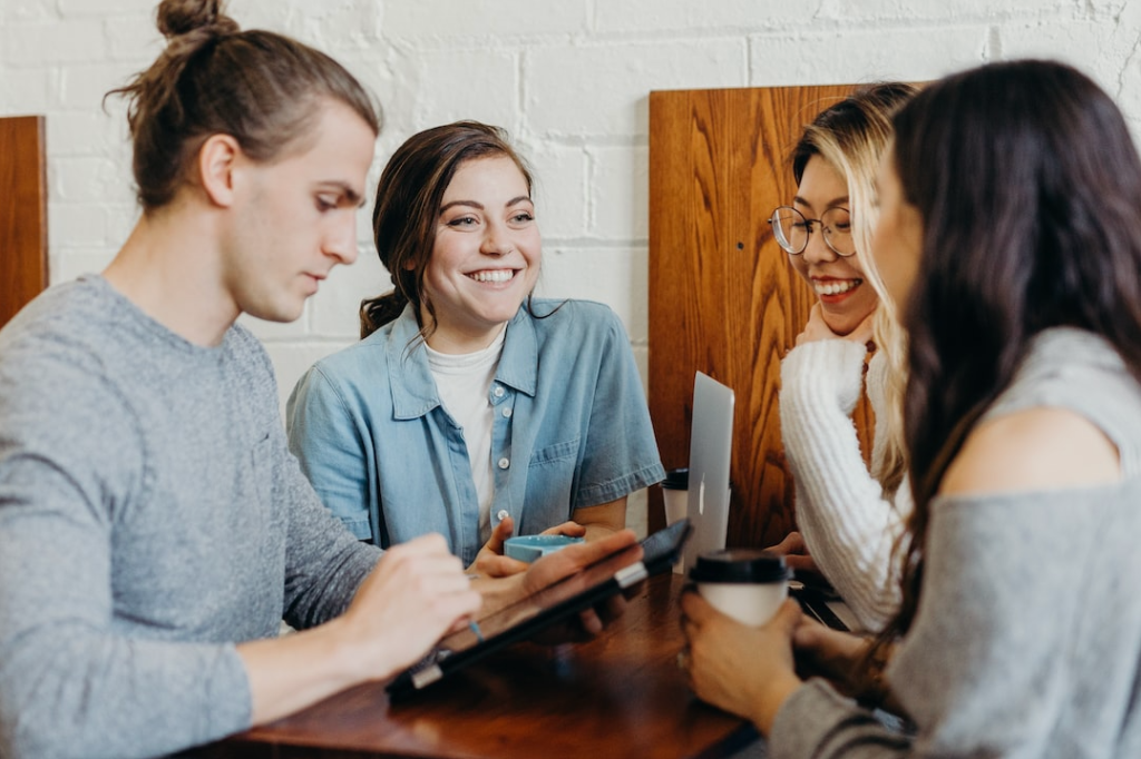 A group of four people sitting at a wooden table, engaged in conversation, and smiling. One person is holding a tablet while another is holding a coffee cup. They are dressed casually, and the background features a white brick wall and wooden panels.