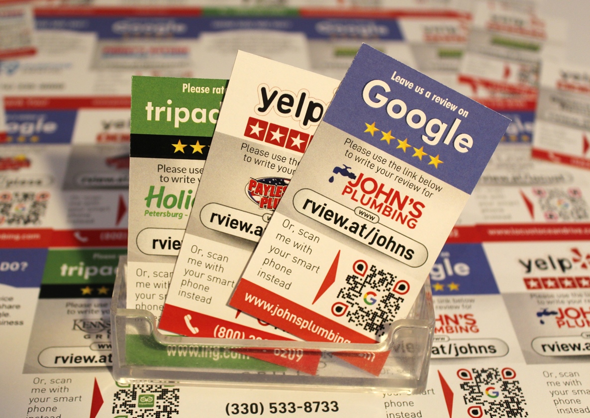 Close-up of a clear holder containing business cards for reviews on Yelp, TripAdvisor, and Google. The cards belong to John's Plumbing and show QR codes and website links for leaving reviews with a prompt: "How do you ask a customer for a review?". Other cards are scattered in the background. Contact number (330) 533-8733.