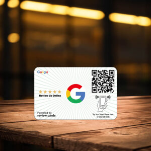 A small Classic Google Smart Review Cards - Free Delivery on a wooden table features the Google logo with a five-star rating above "Review Us Online." There's a QR code on the right and an NFC tag illustration below it. The background is blurry, showing warm lights and hints of an indoor setting.