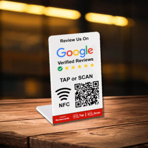 A display stand on a wooden table encourages customers to review a business on Google Verified Reviews by tapping or scanning a QR code. The card highlights NFC technology and has a web address labeled "review.cards." The background is a warmly lit, blurred interior, showcasing 1 Google Tap or Scan Stands - FREE Delivery.
