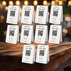 Several white 10 Google Tap or Scan Table Tents are placed on a wooden surface, each displaying a “Review Us On Google” header, a QR code, star rating visuals, and a space for comments. The backdrop is a cozy, dimly-lit setting with hanging lights in soft focus.