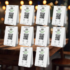 A wooden table in a restaurant displays several identical 10 TripAdvisor Tap or Scan Table Tents encouraging reviews. Each card features the TripAdvisor logo, a QR code, and the message "Review us on TripAdvisor." The background shows more tables slightly out of focus.