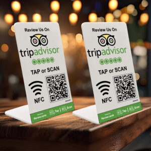 Two 2 TripAdvisor Tap or Scan PoP Stands inc FREE Delivery on a wooden surface encourage patrons to review on TripAdvisor. Each stand displays the TripAdvisor logo and features instructions to "TAP or SCAN" using NFC or QR codes for easy access. Blurred lights create a cozy, inviting background atmosphere.