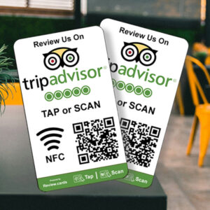 Two "2 TripAdvisor Tap or Scan Smart Review Cards - Free Delivery" are shown, featuring the TripAdvisor logo at the top. Each card includes a QR code, NFC symbol, and instructions to "Tap or Scan on TripAdvisor." The cards are positioned on a table, with a blurred café background and yellow chairs.