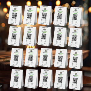 An array of folded 20 TripAdvisor Tap or Scan Table Tents displayed in a grid formation, each featuring the TripAdvisor logo and QR codes for customer reviews. The background is a warmly lit, blurry setting, possibly a restaurant or cafe.