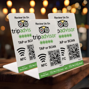 Three 3 TripAdvisor Tap or Scan PoP Stands inc FREE Delivery are displayed on a wooden surface. Each stand features the TripAdvisor logo, icons for NFC and QR code scanning, and the instructions "TAP or SCAN." Blurred, warm background lights create a cozy ambiance.