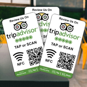 Three rectangular signs with the TripAdvisor logo and text "Review Us On TripAdvisor" are shown. Each sign features a QR code and NFC symbols, along with the instruction to "Tap or Scan." A blurred outdoor setting with a yellow chair is in the background. The product displayed is 3 TripAdvisor Tap or Scan Smart Review Cards - Free Delivery.