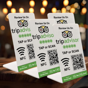 Four small triangular 4 TripAdvisor Tap or Scan PoP Stands inc FREE Delivery rest on a wooden table, each displaying "Review Us On tripadvisor" with the TripAdvisor logo, a QR code, and NFC symbol. Blurred warm lights in the background create a cozy atmosphere.
