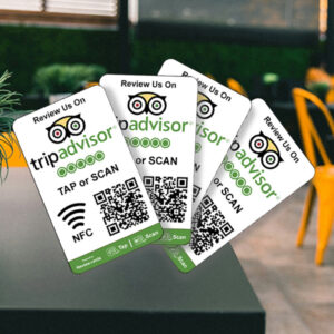 Four 4 TripAdvisor Tap or Scan Smart Review Cards are fanned out on a table, each promoting users to Tap or Scan with TripAdvisor for easy reviewing. The cards feature NFC and QR code options amidst a blurred indoor setting with plant decor and yellow chairs.