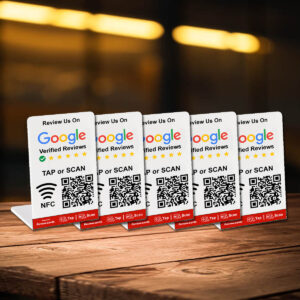 Five tabletop stands display cards that prompt customers to review a business on Google. Each card shows a QR code and NFC symbol for easy access, with instructions to "TAP or SCAN." The background is out of focus, suggesting a cozy indoor setting. These 2 Google Tap or Scan Stands - FREE Delivery make reviewing effortless.