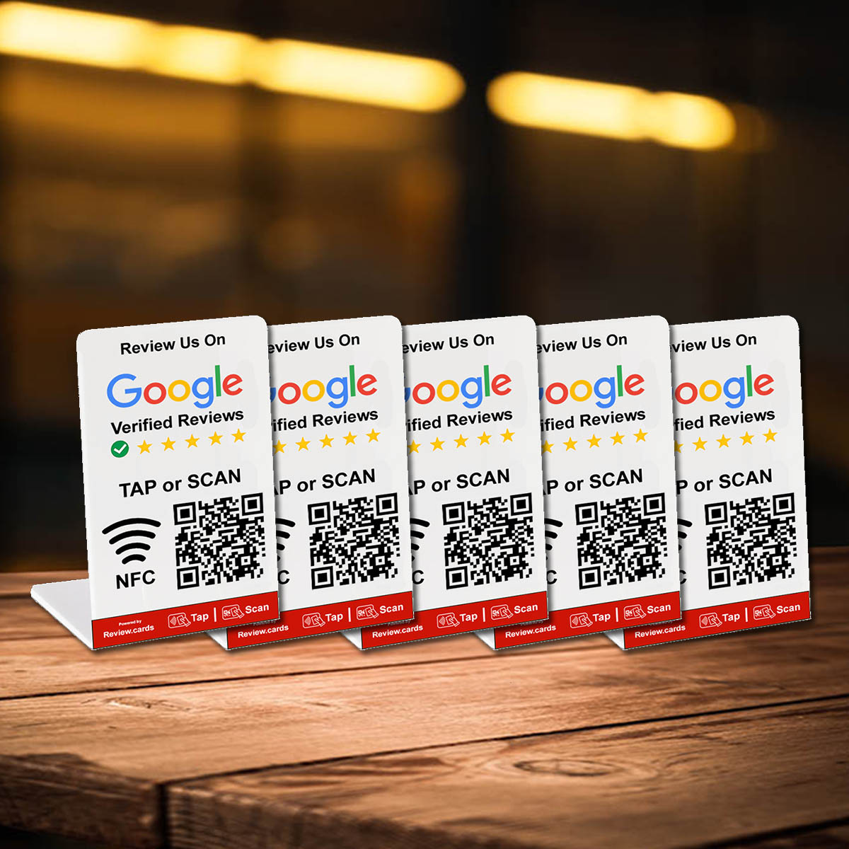 Five tabletop stands display cards that prompt customers to review a business on Google. Each card shows a QR code and NFC symbol for easy access, with instructions to "TAP or SCAN." The background is out of focus, suggesting a cozy indoor setting. These 2 Google Tap or Scan Stands - FREE Delivery make reviewing effortless.