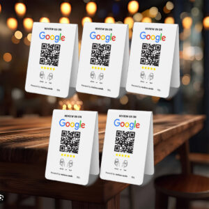Five 5 Google Tap or Scan Table Tents with QR codes on a wooden table, each displaying the Google logo and the text "REVIEW US ON Google," along with illustrations of thumbs up and five stars. The background features warm, blurred bokeh lights, suggesting a cozy, inviting atmosphere.
