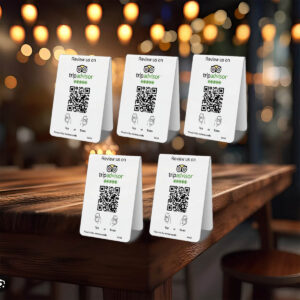 A wooden table in a dimly lit restaurant displays five 5 TripAdvisor Tap or Scan Table Tents with QR codes inviting customers to review the establishment on TripAdvisor. The background features bokeh lights, creating a warm and cozy atmosphere. Simply tap or scan to share your experience!