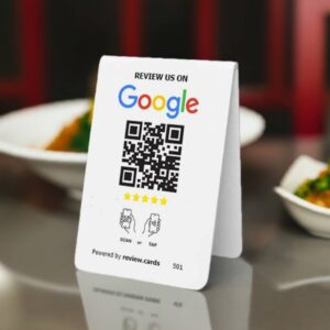 A Google Tap or Scan Table Tent encourages customers to review the establishment on Google. The card, set on a table, shows a 5-star rating graphic and instructions to "Scan or Tap." Plates of food are slightly blurred in the background.