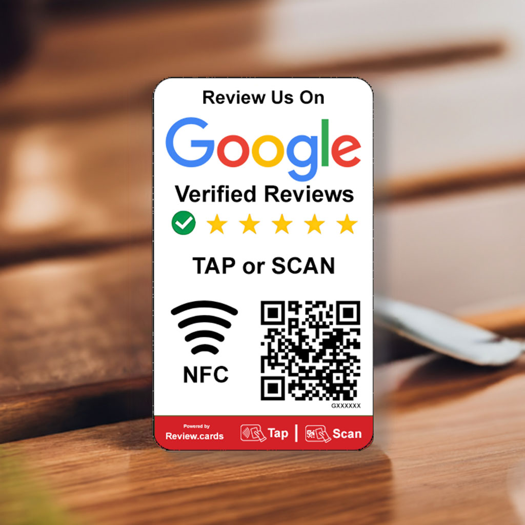 A sign prompting customers to review on Google features the Google logo, five stars, and the text "Verified Reviews." It invites users to "TAP or SCAN" with NFC and QR code options. The background shows a blurred wooden surface, possibly a table.