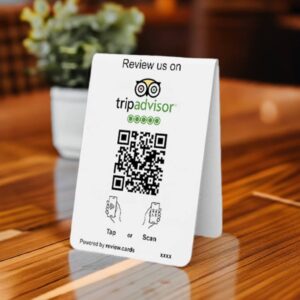 A TripAdvisor Tap or Scan Table Tent on a wooden table encourages patrons to tap or scan the QR code to review the business. It features the TripAdvisor logo and clear instructions. A potted plant and a blurred background enhance the inviting setting.